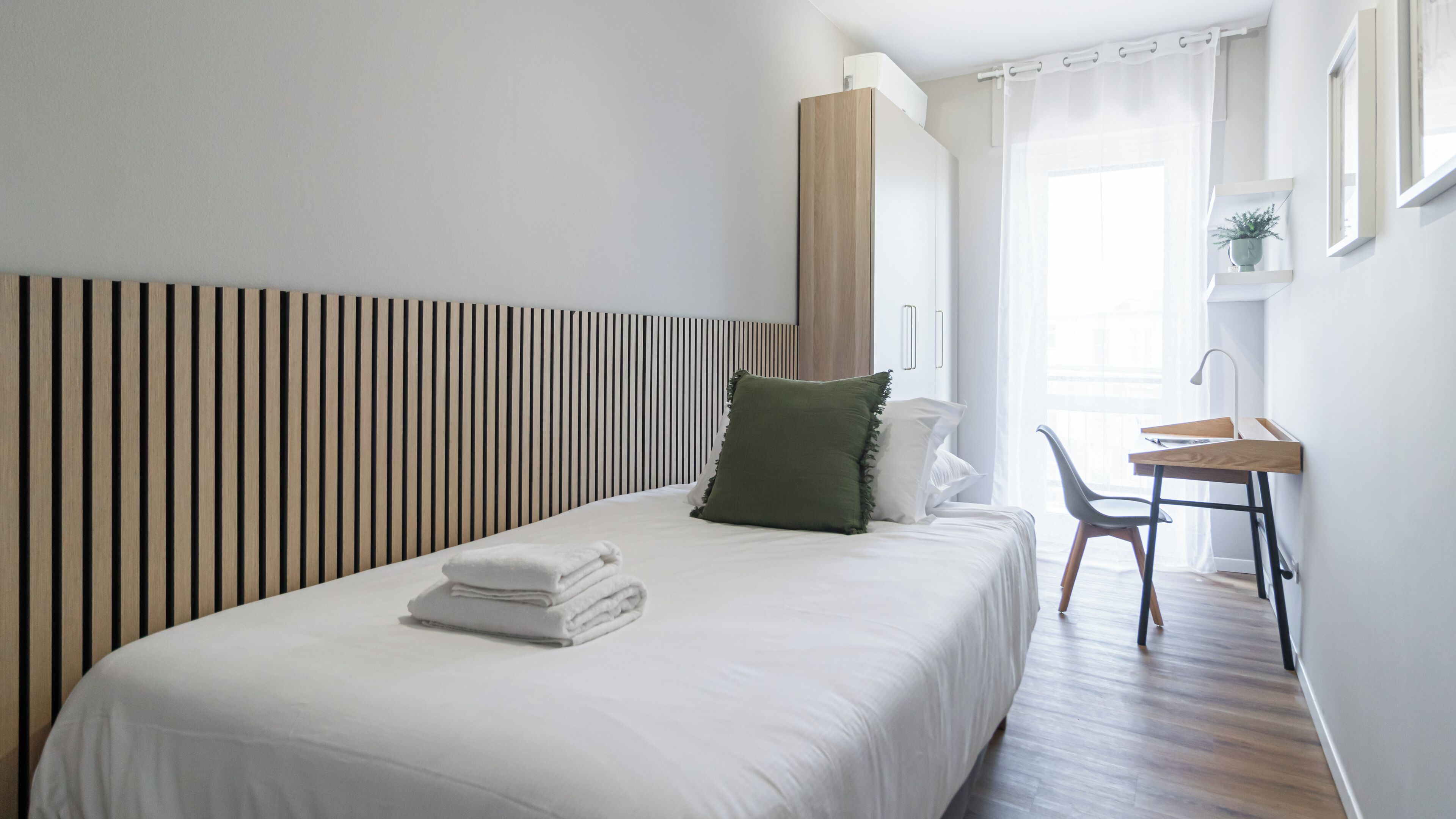 A neatly made bed with white linens and a green pillow, a wooden desk with a chair, and a window with sheer curtains.