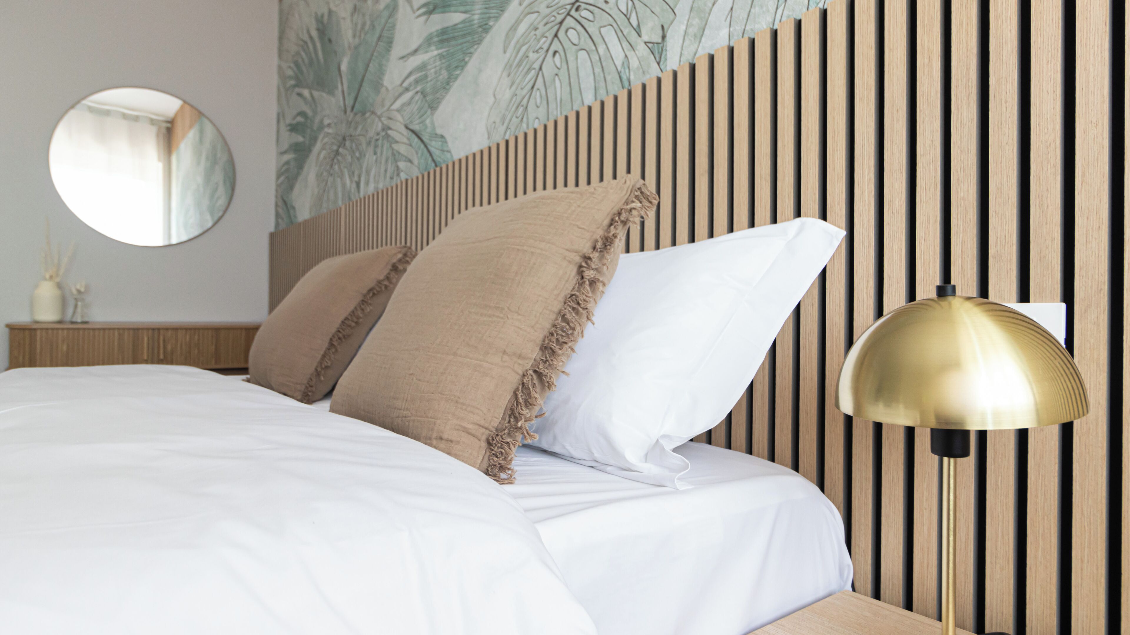 A neatly made bed with white linens and two pillows, a round mirror on the wall, and a wall-mounted lamp.