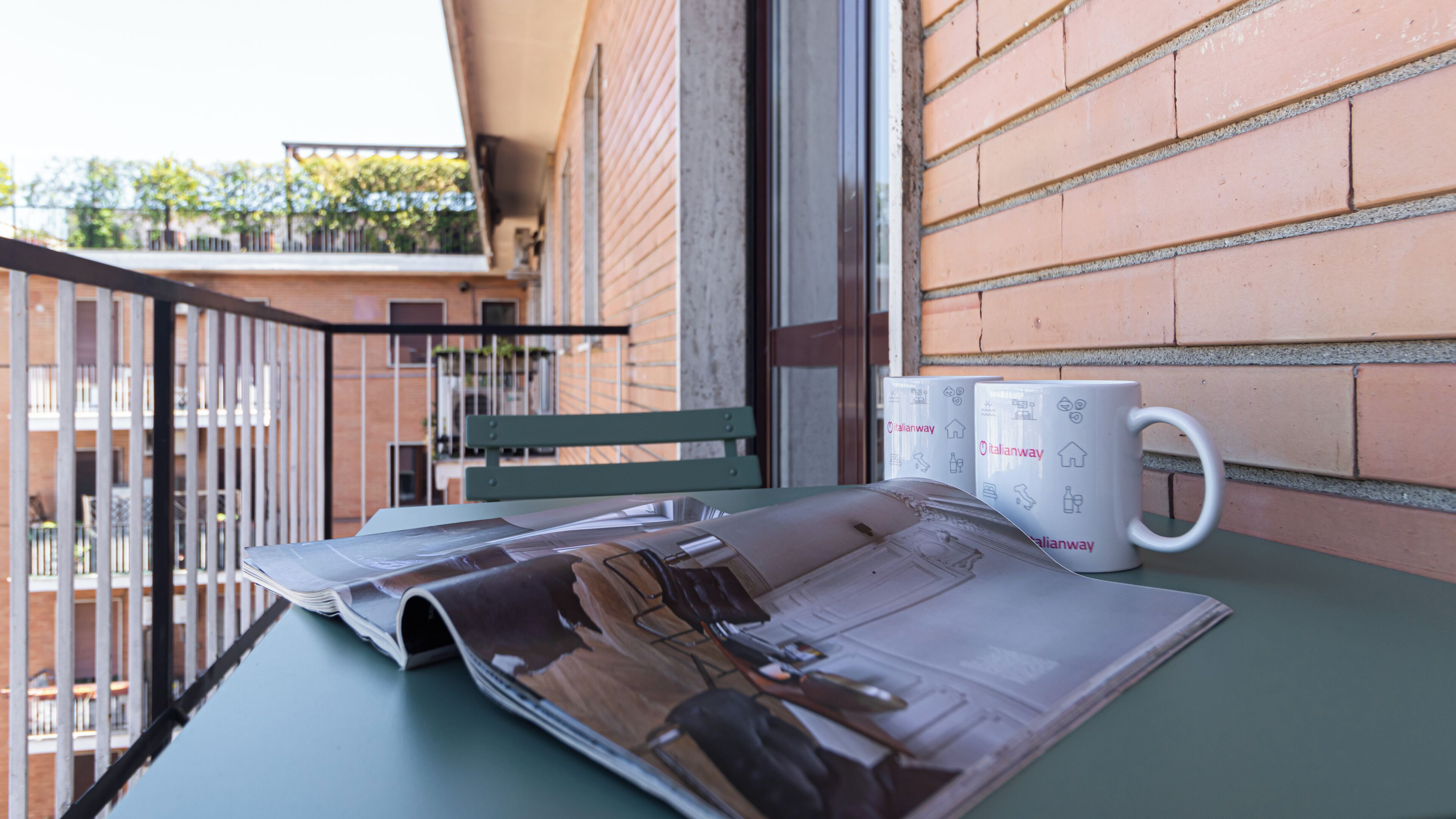 A balcony with a table, two white mugs with Italian symbols, and a green chair.
