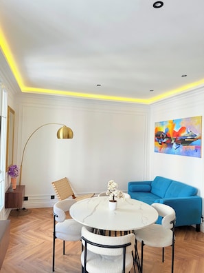 A modern living room with a round table, white chairs, a blue sofa, a floor lamp, and a colorful painting on the wall.