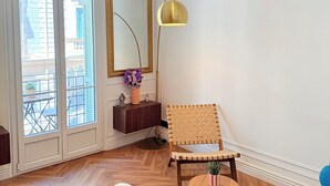 A room with a large window, a mirror, a floor lamp, a chair, a small table, a blue sofa, and a white ottoman. - Charming 2-room apartment with balcony (Nice)