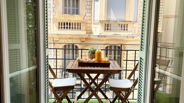 A room with a sliding glass door leading to a balcony with a table and chairs, a small potted plant, and a bottle of orange juice. - Charming 2-room apartment with balcony (Nice)