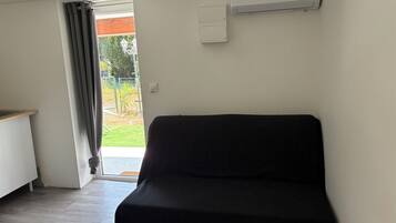 A room with a black sofa, a wooden table, a white air conditioning unit, and a view of a garden through a door.
