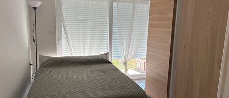 Et lille soveværelse med en seng, et natbord, en lampe og et vindue med gardiner.
