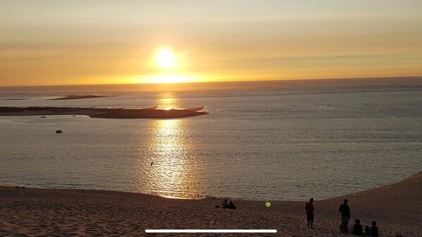 Une plage au coucher du soleil, avec le soleil bas à l’horizon, une mer calme et des gens sur le sable.