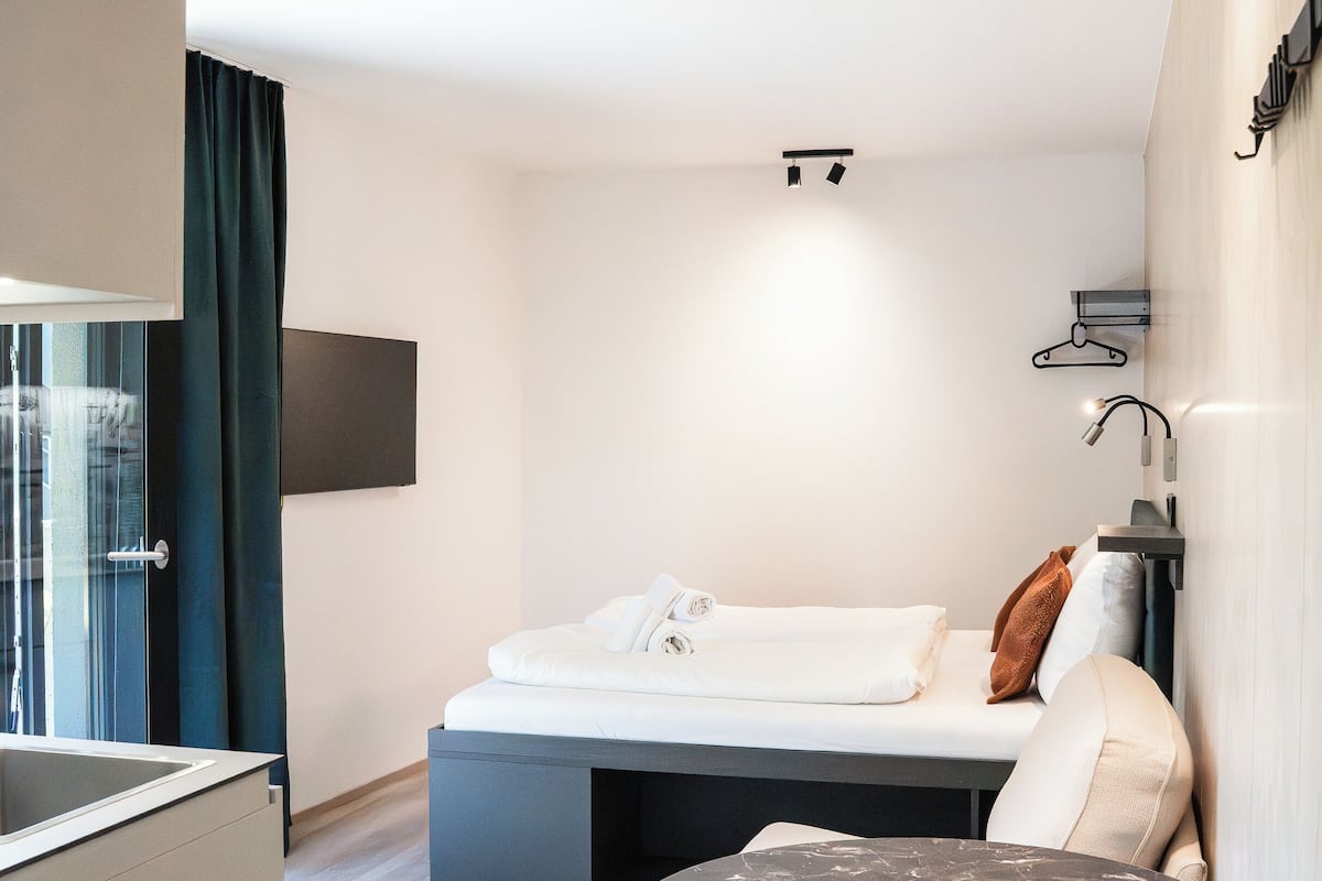 A modern hotel room with a bed, a small round table, a sofa, a TV mounted on the wall, and a window with curtains.
