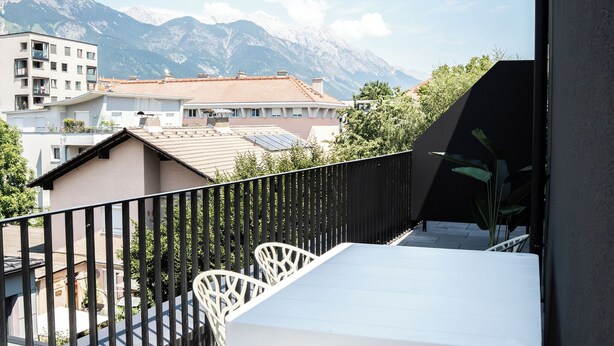 A balcony with a white table and chairs, a black railing, and a view of buildings and mountains in the background.