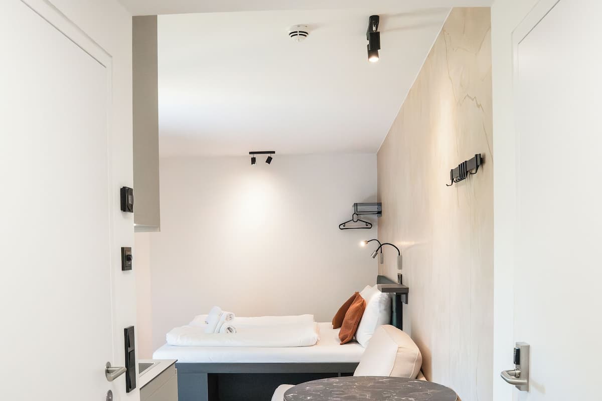 A modern hotel room with a minimalist design, featuring a bed, a small round table, and a red ottoman.