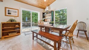 Un coin repas avec une table en bois, des chaises et un banc. On y trouve une bibliothèque remplie de livres et d’objets décoratifs. La pièce est dotée d’un plafond en bois et de grandes fenêtres offrant une vue sur la verdure environnante.