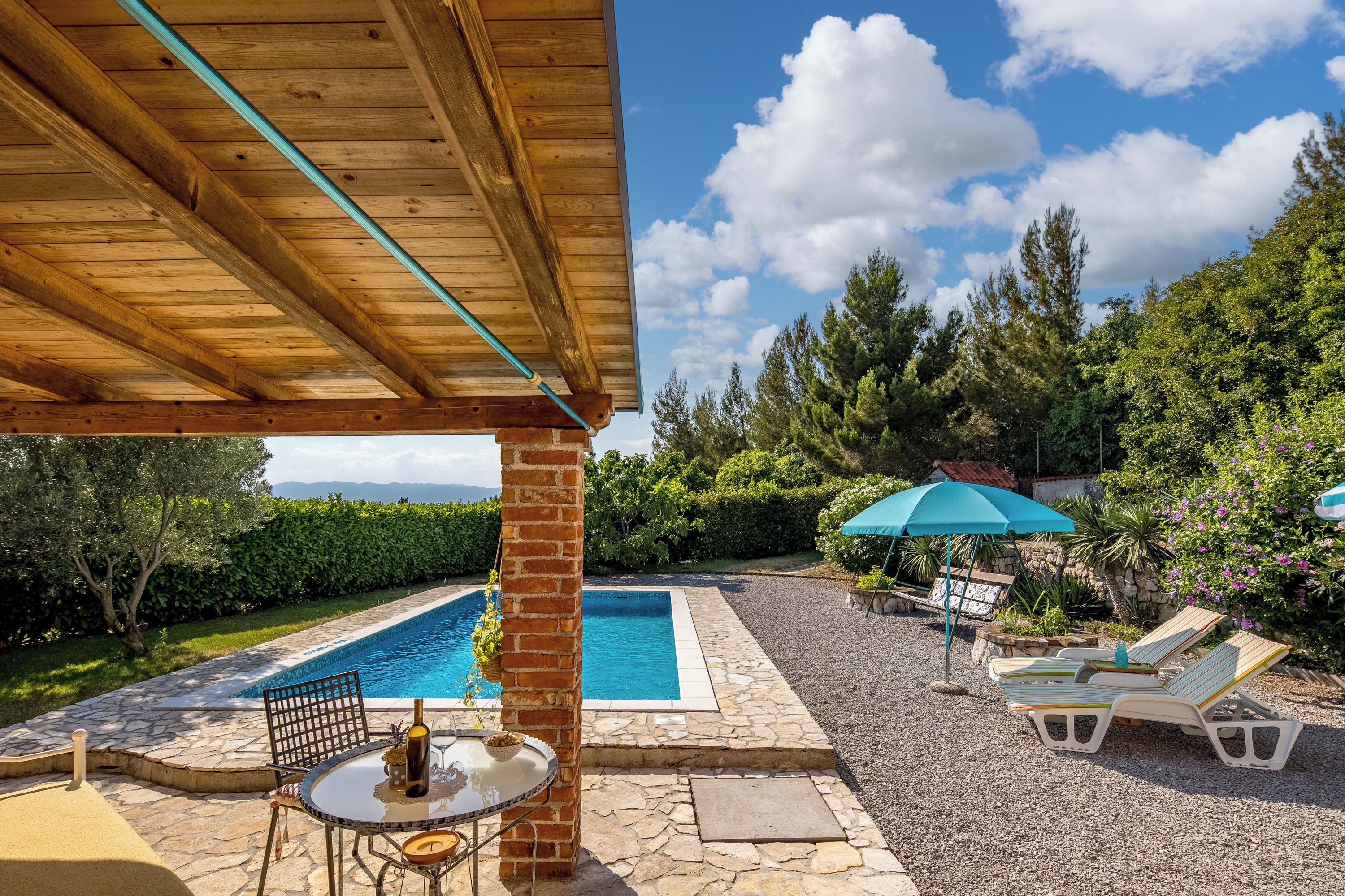 A pool area with lounge chairs and a table, surrounded by greenery and a clear sky.