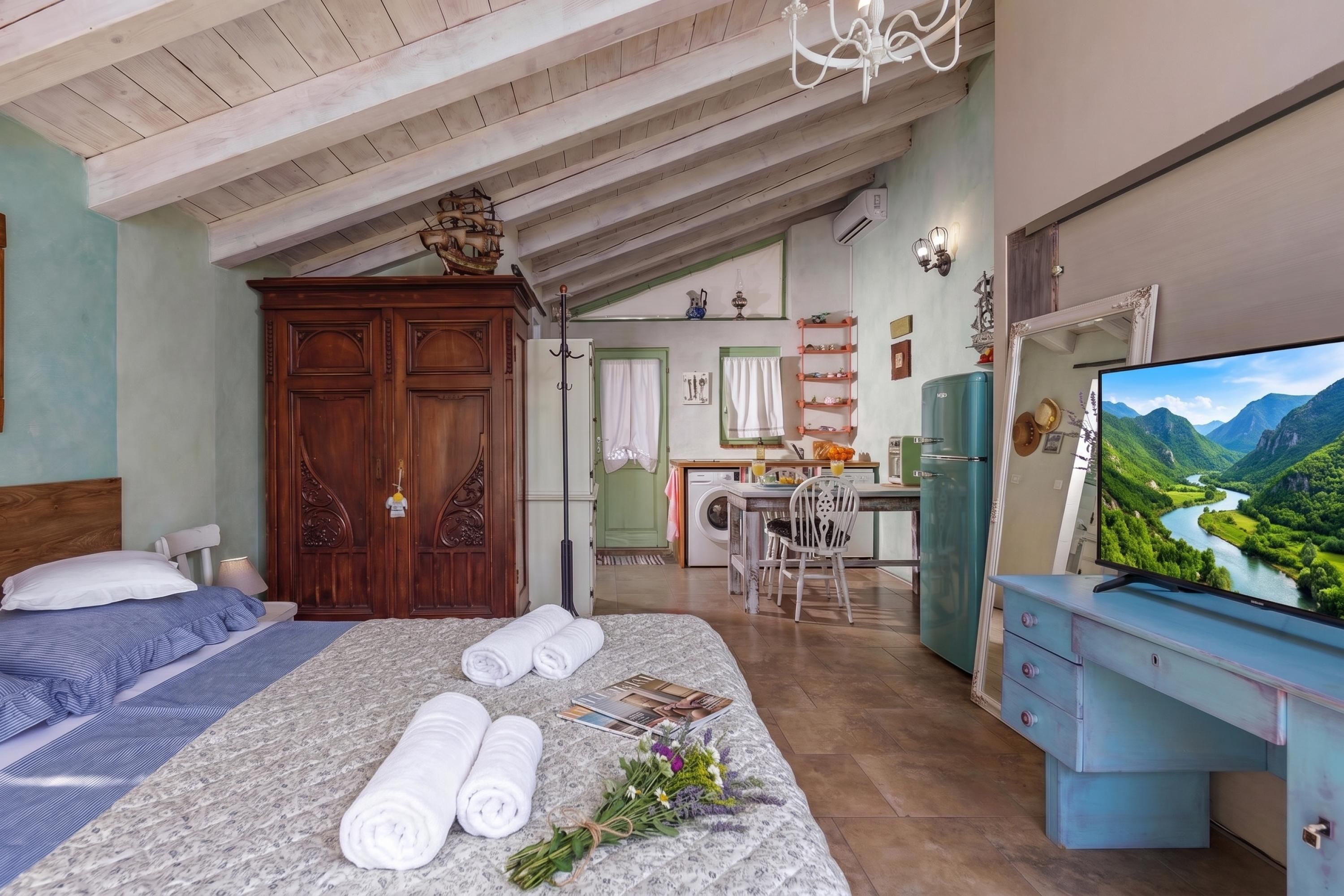 Un dormitorio con cama, armario de madera, zona de comedor con mesa y sillas, cocina con refrigerador y televisor que muestra una vista panorámica.