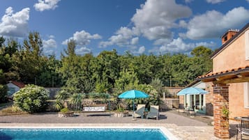 Una piscina con sillas de descanso y una sombrilla, rodeada de vegetación y un muro de piedra.