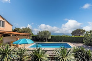 Une piscine avec un parasol bleu, entourée de verdure et offrant une vue sur la mer.