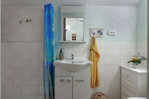 Une salle de bain avec un lavabo blanc, un miroir et un meuble. On y trouve un rideau de douche, une serviette et une petite plante sur le plan de toilette.