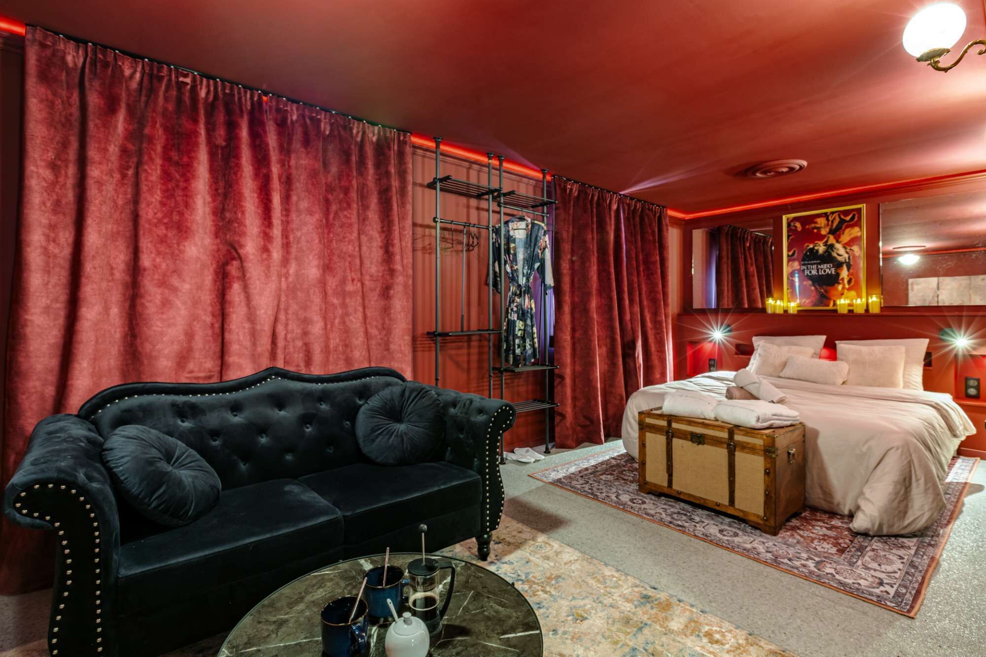 Une pièce avec un canapé noir, un lit avec du linge de lit blanc, une commode en bois et un rideau rouge.