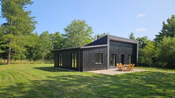 A modern wooden cabin with large windows, a patio with outdoor furniture, surrounded by a grassy field and trees. - Létendart (Saint-Quentin-en-Tourmont)