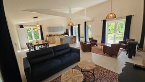 A spacious living area with a black sofa, two round coffee tables, and a patterned rug. A kitchen with light wood cabinets and a dining area with a table and chairs are visible in the background. - Létendart (Saint-Quentin-en-Tourmont)