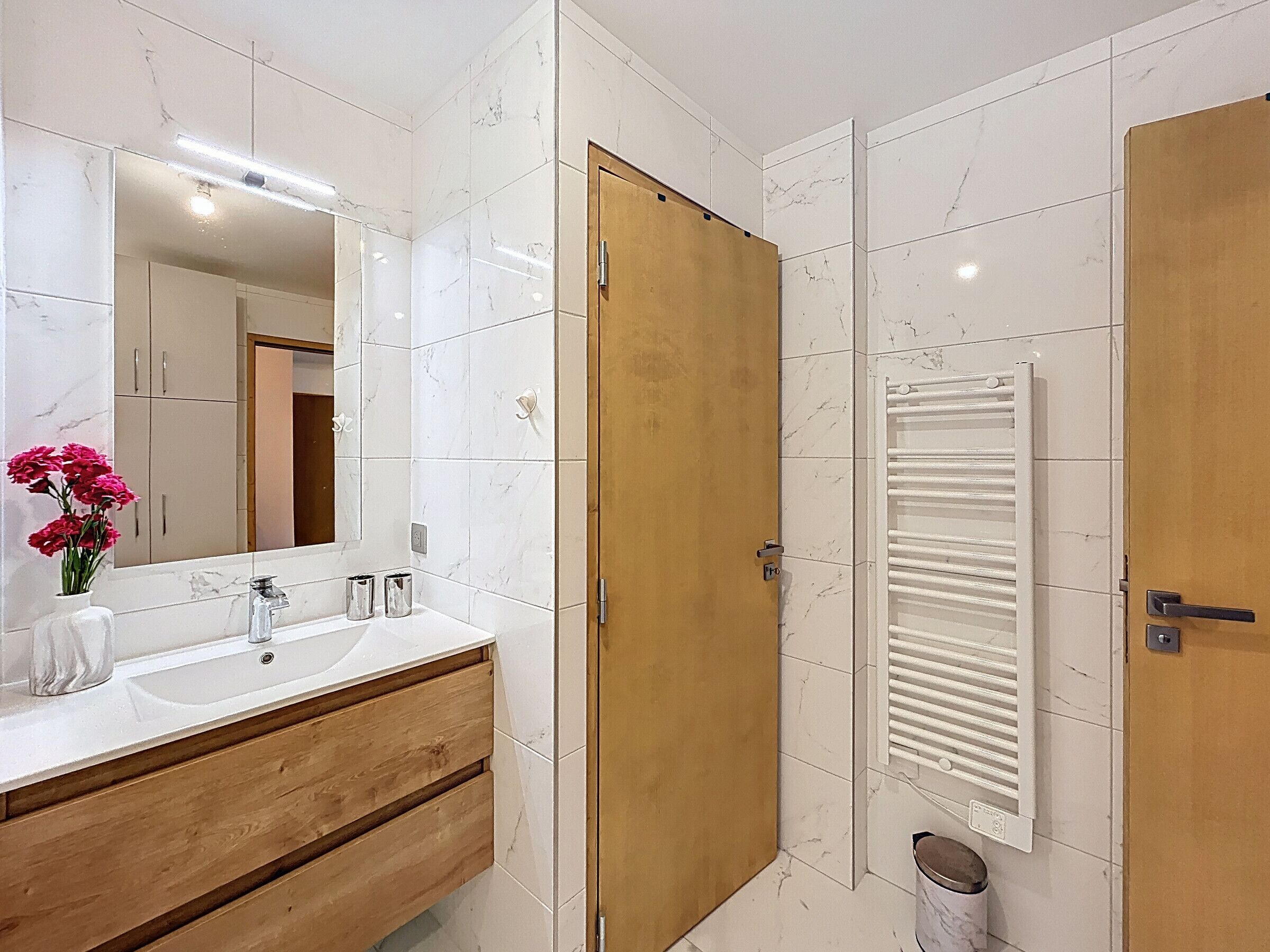 A modern bathroom with a wooden vanity, a large mirror, and a white tiled wall.