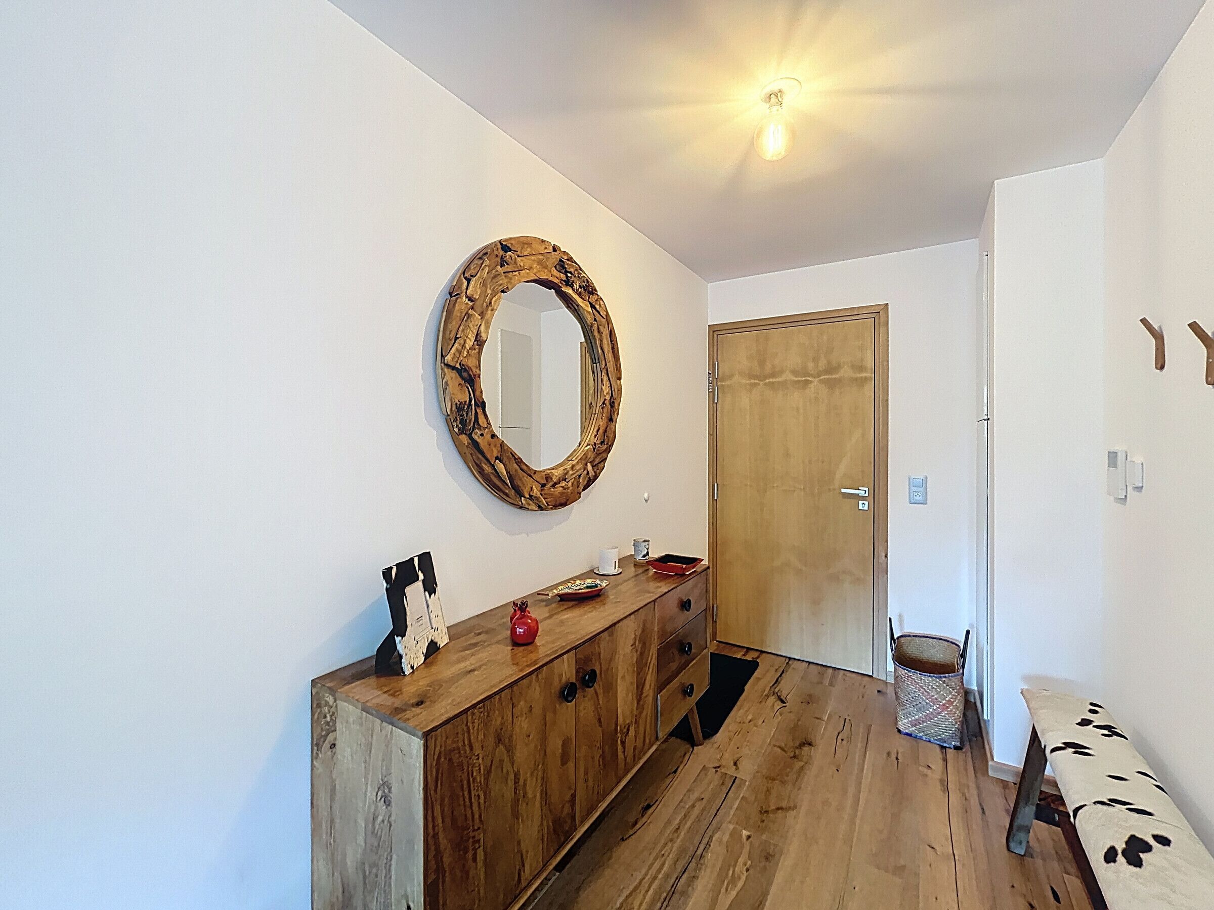 A room with a wooden cabinet, a round mirror, a door, and a bench.