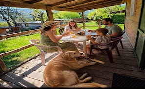Un espace extérieur abrité, aménagé pour les repas, avec une terrasse en bois, une table et des chaises, et un chien allongé sur le sol.