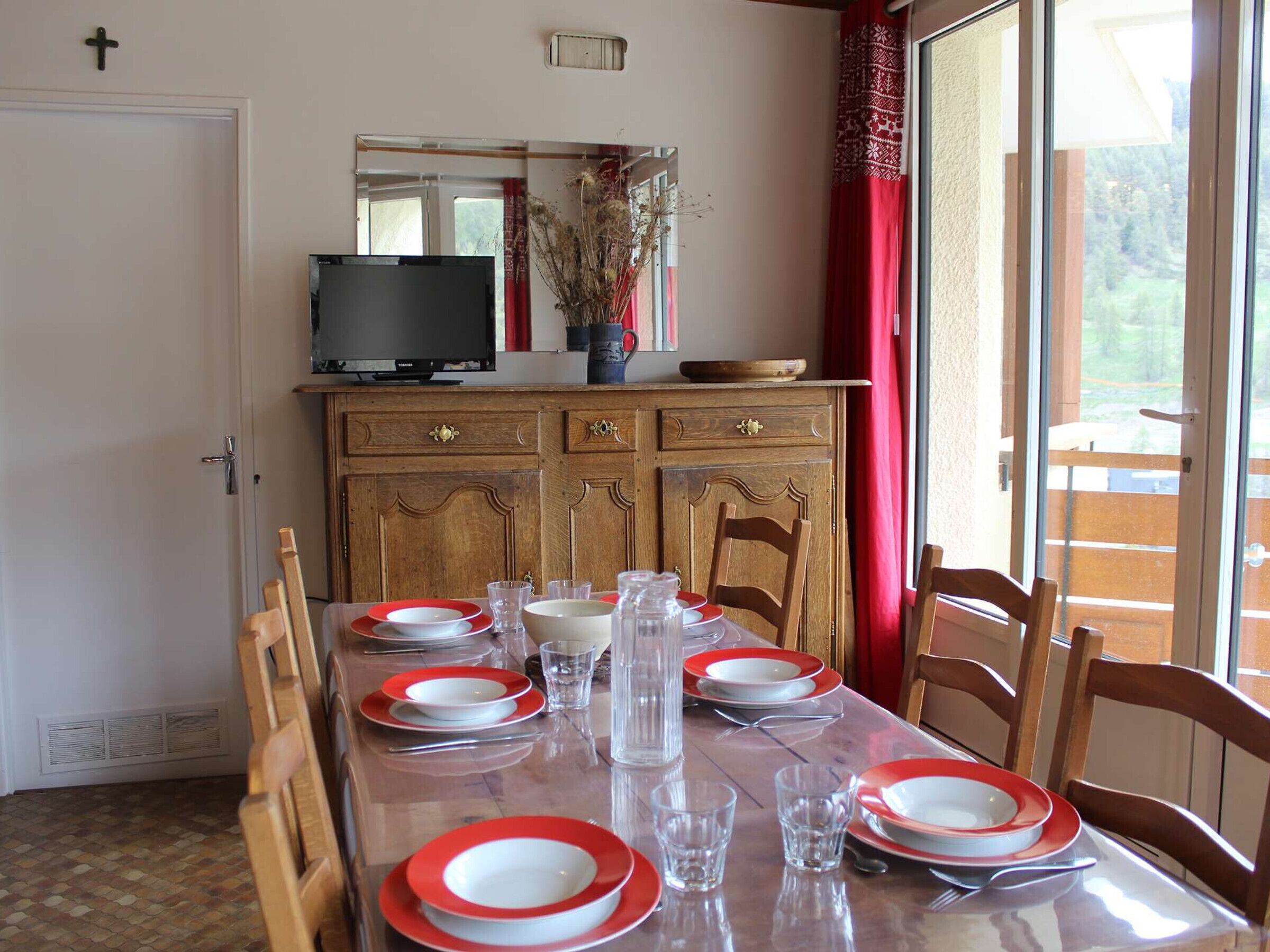 A dining room with a wooden table set for four, a television on a cabinet, and a window with red curtains.