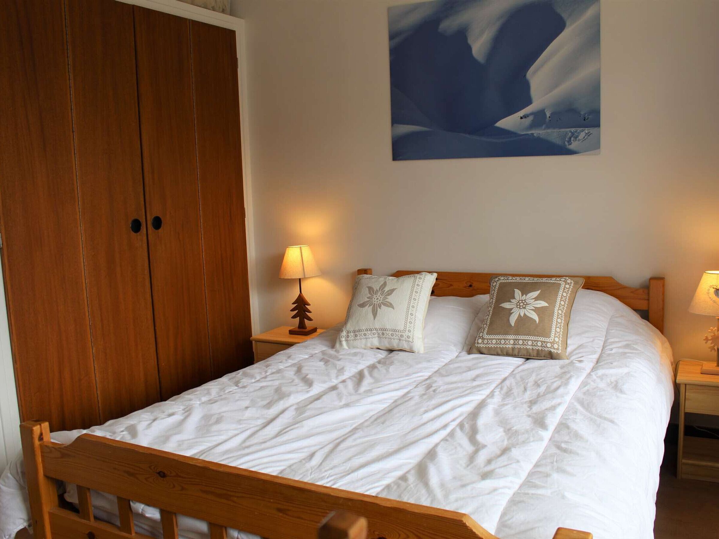 A bedroom with a wooden bed frame, white bedding, two bedside lamps, and a large painting of a snowy landscape.