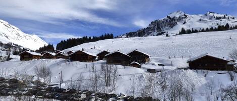 A snow-covered mountain village with wooden cabins, a ski slope, and a clear blue sky.