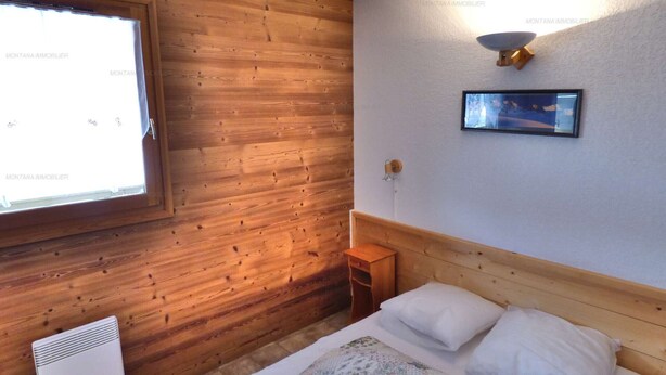 A room with wooden panel walls, a bed with white bedding, a small bedside table, a wall-mounted light fixture, and a window with curtains.