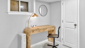 Habitación pequeña con un escritorio de madera, una silla, una lámpara de escritorio, un reloj, un radiador, un espejo y una puerta.