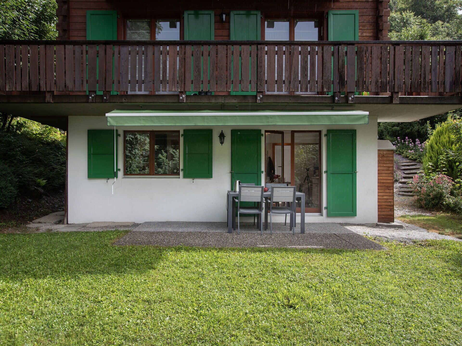 Una casa de dos pisos con contraventanas verdes, un balcón y un pequeño patio con mesa y sillas.