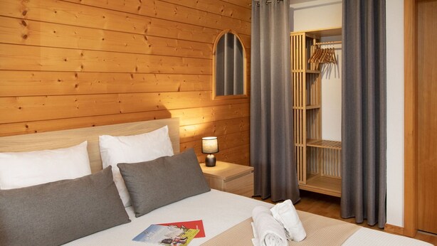 A hotel room with a wooden headboard, a bed with white and gray bedding, a nightstand with a lamp, a towel, and a magazine on the bed. - Lovely apartment for 3 guests with WIFI, TV and terrace (Ovronnaz)