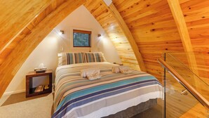 Un dormitorio en el ático con techo de madera inclinado, una cama con ropa de cama a rayas, una mesita de noche pequeña y una ventana con persianas.