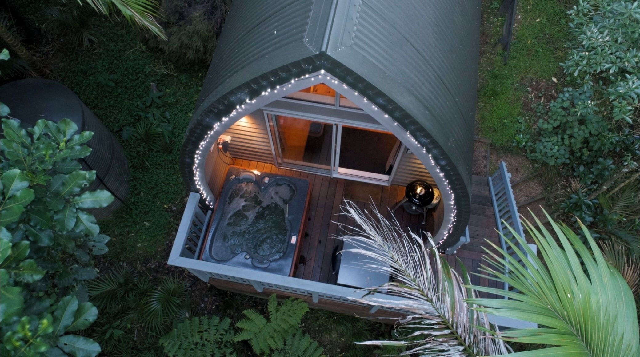 A hot tub with a wooden deck and a small seating area, surrounded by greenery.