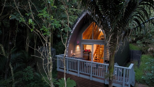 Una casa en el árbol con terraza de madera y acceso de vidrio, rodeada de follaje denso.