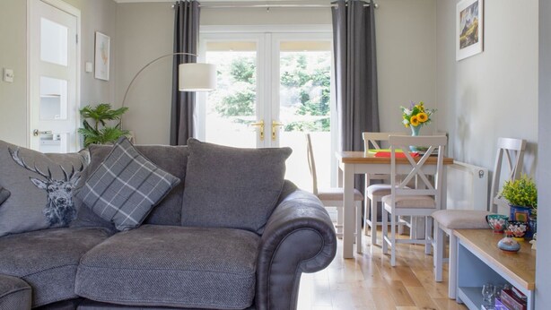 A modern living room with a grey sofa, a dining table with chairs, a bookshelf, and a large window with curtains. - Beautiful  villa for 5 guests with WIFI and TV (Isle Of Skye)