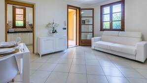 Ein geräumiges Wohnzimmer mit einem weißen Sofa, einem Esstisch mit Tellern und einem Glas sowie einer weißen Kommode mit einem Blumenstrauß.