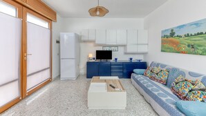 Ein kompakter Wohnbereich mit Küchenzeile, einer Couch mit gemusterten Kissen, einem Couchtisch und einem Bild an der Wand.