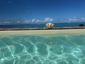 Una piscina cristal·lina amb un pont de fusta i una gran decoració de closca, amb vista a una platja i l'oceà.