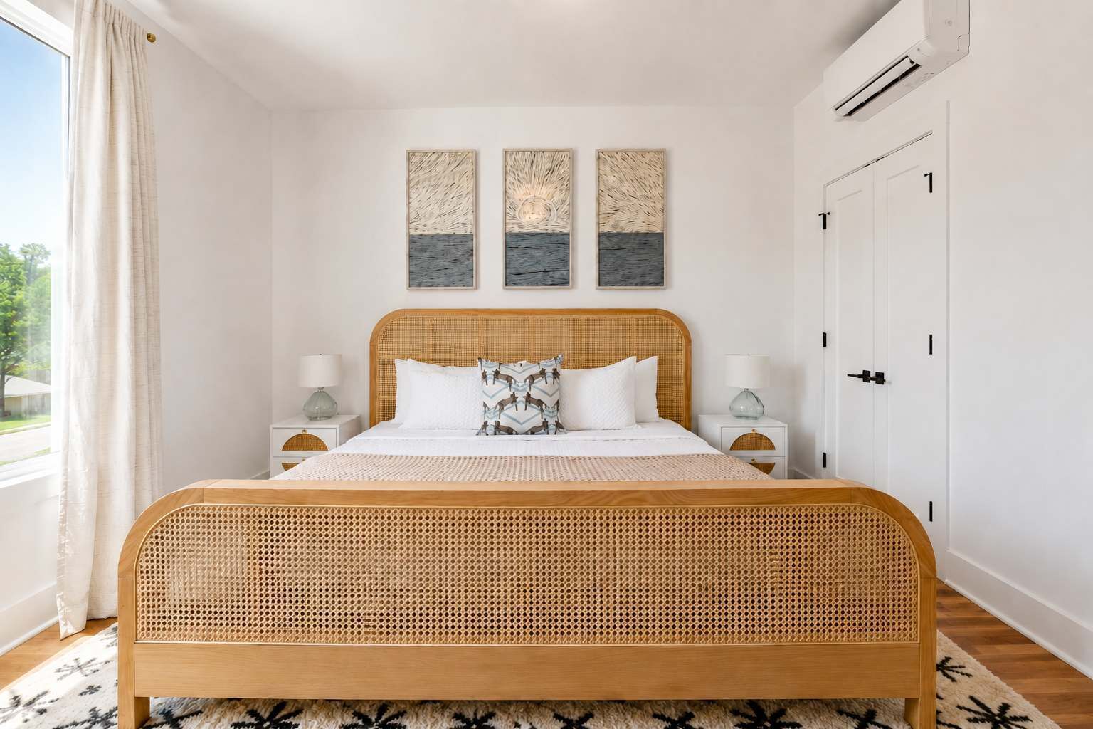 Een slaapkamer met een rieten bed, twee nachtkastjes met lampen, een raam met gordijnen en drie aan de muur bevestigde kunstwerken.