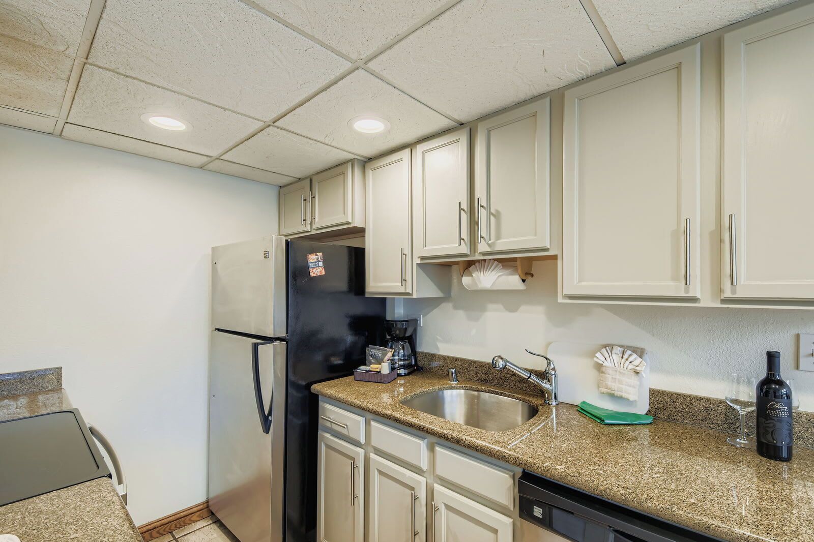 A kitchen with white cabinets, a stainless steel refrigerator, a coffee maker, a sink, and a wine bottle.