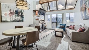 A spacious living area with a large table, comfortable seating, a fireplace, and a view of a snowy landscape through large windows.