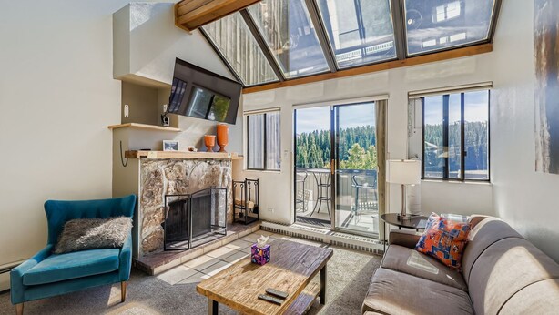 A spacious living room with a stone fireplace, a TV mounted on the wall, and a balcony with a scenic view.