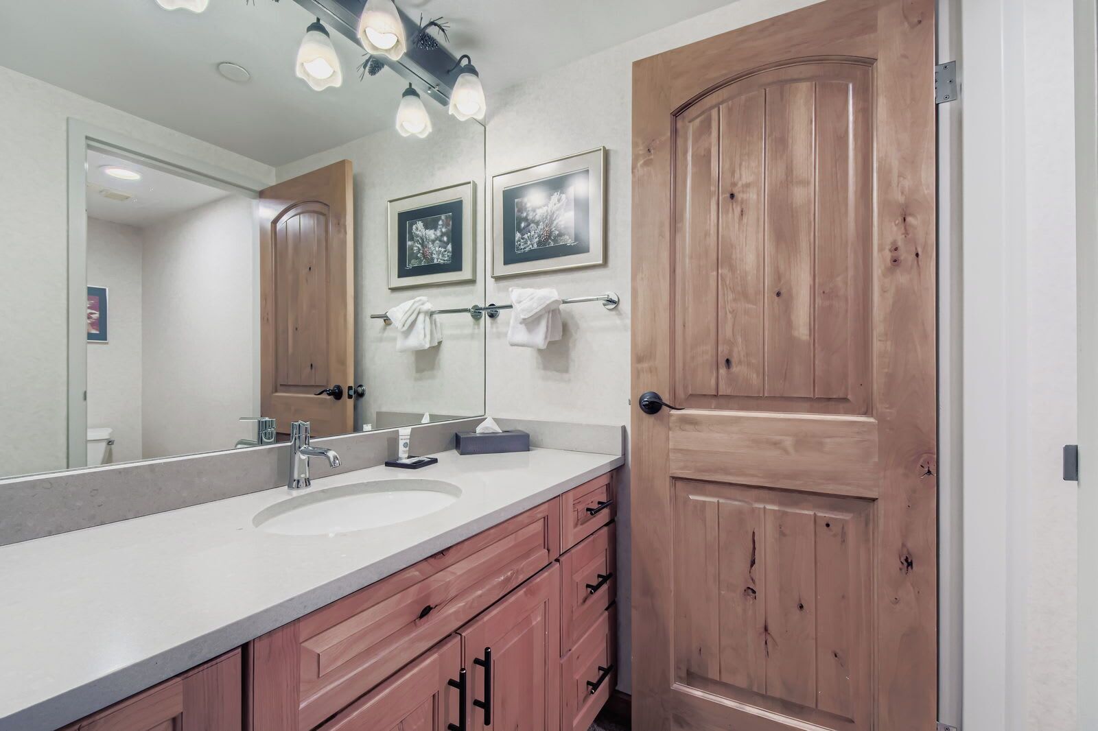 A bathroom with a double sink, wooden cabinets, a large mirror, and a wooden door.