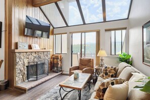 A spacious living room with a stone fireplace, a comfortable sofa, a television mounted on the wall, and large windows offering a view of the outdoors.