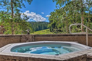 A hot tub with a stone wall and metal railing, surrounded by trees and a view of a green hillside under a clear blue sky.