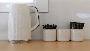 Una tetera blanca con un asa texturizado, junto a tres recipientes blancos etiquetados 'SUGAR', 'TEA' y 'COFFEE'.