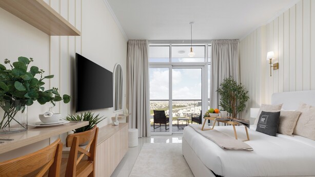 A modern hotel room with a flat-screen TV, wooden furniture, a sofa, a dining area, and a balcony with a view. - Maison Privee - Stylish Studio w/ Top Facilities in Damac Hills (Dubai)