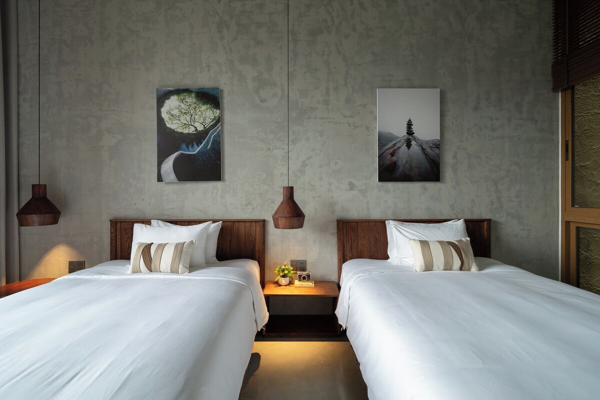 Habitación de hotel con dos camas, una mesita de noche con lámpara y dos obras de arte colgadas en la pared.
