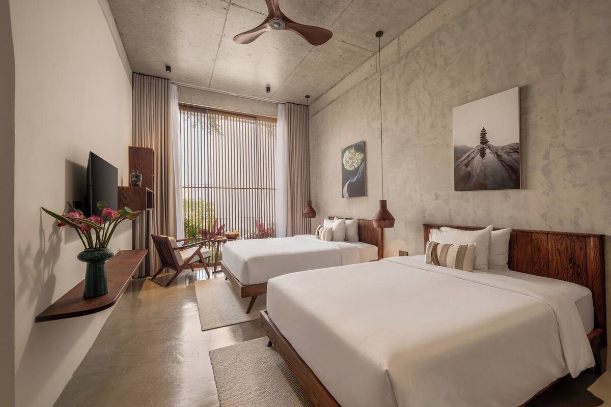 Une chambre d’hôtel avec deux lits, un ventilateur de plafond, une grande fenêtre avec des rideaux, un téléviseur à écran plat et des œuvres d’art accrochées aux murs.
