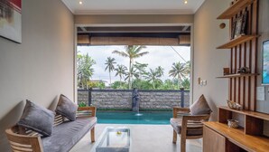 A modern living room with a glass coffee table, wooden furniture, and a view of a swimming pool with palm trees. - Alisa Private Villa (Ubud)